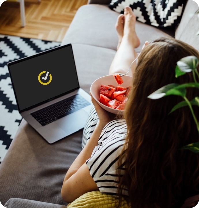 Femme allongée sur un canapé, mangeant un bol de fruits, avec un ordinateur portable à côté d'elle affichant le logo Norton